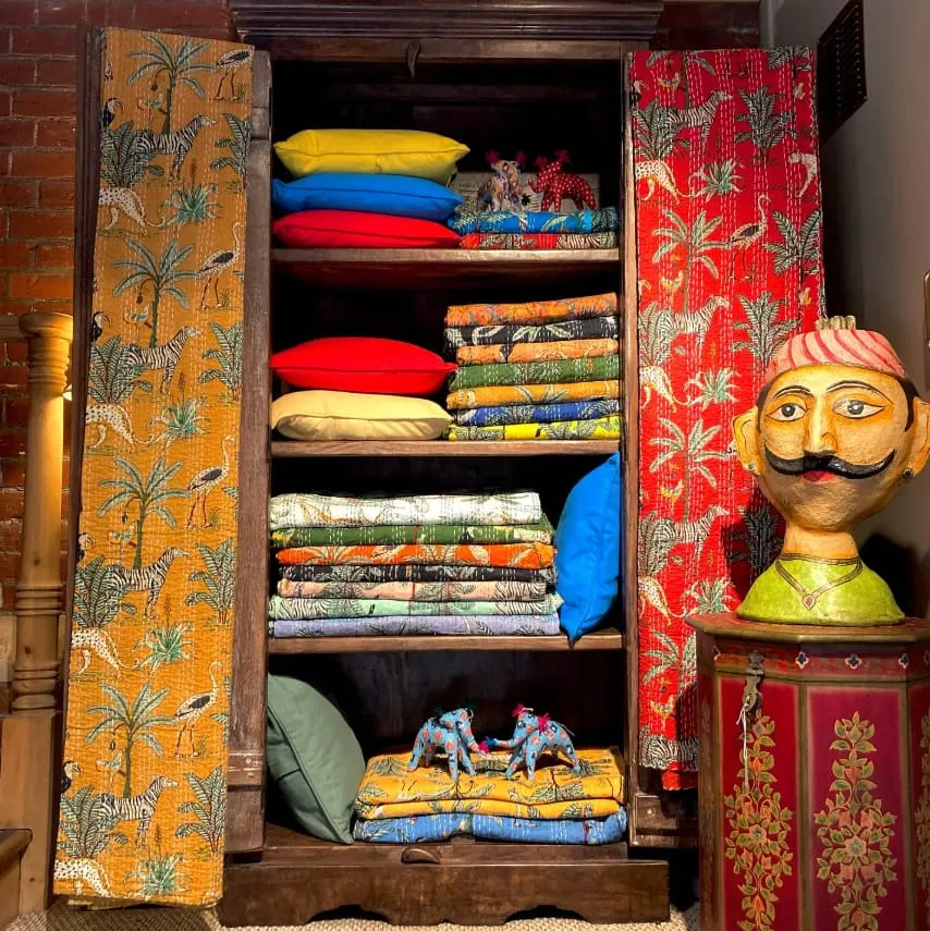 Wooden cabinet filled with neatly stacked colourful cushions and fabrics with patterned doors, alongside a decorative wooden head sculpture.