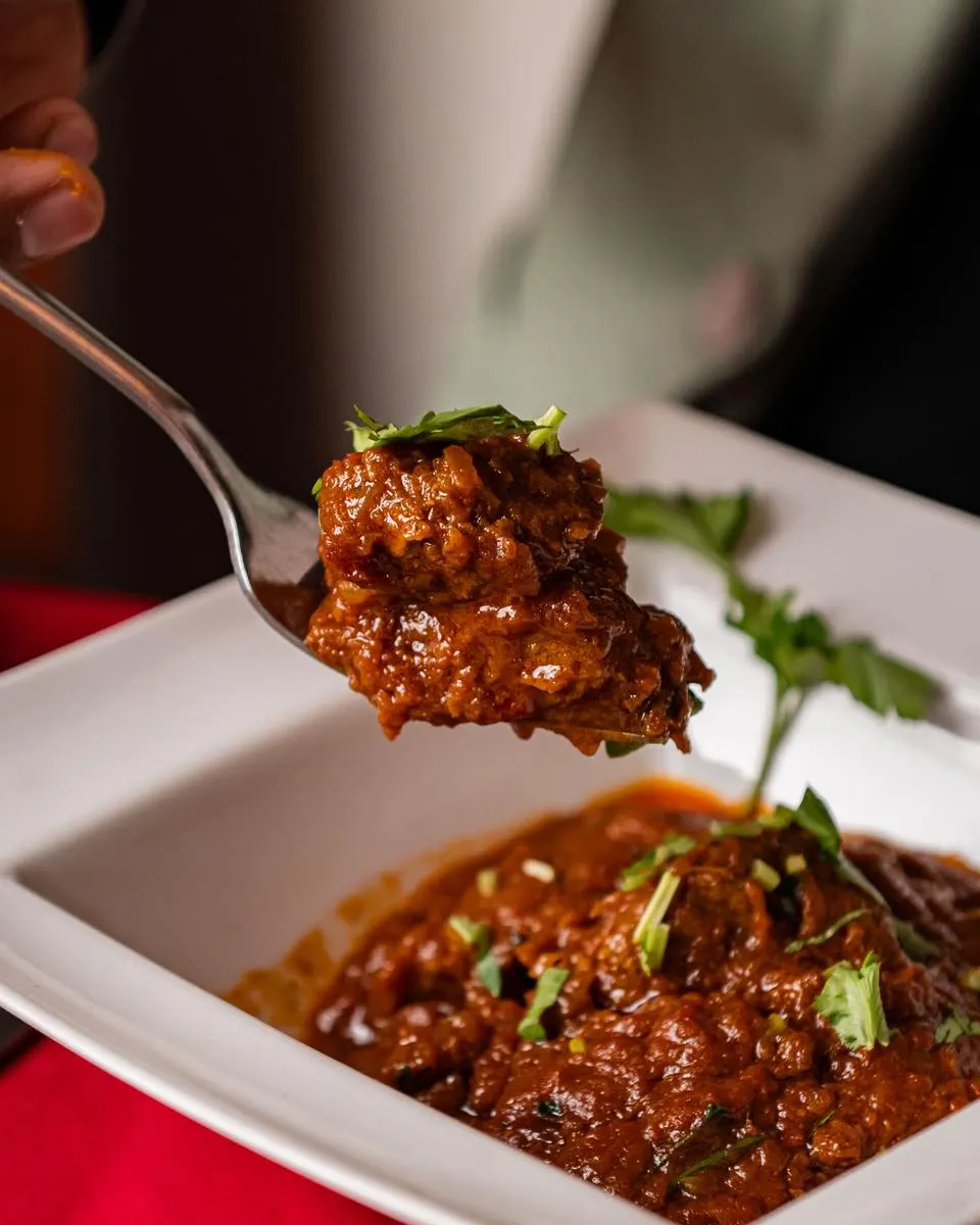 A forkful of spiced meat curry held above a white bowl of the same dish, garnished with fresh coriander.
