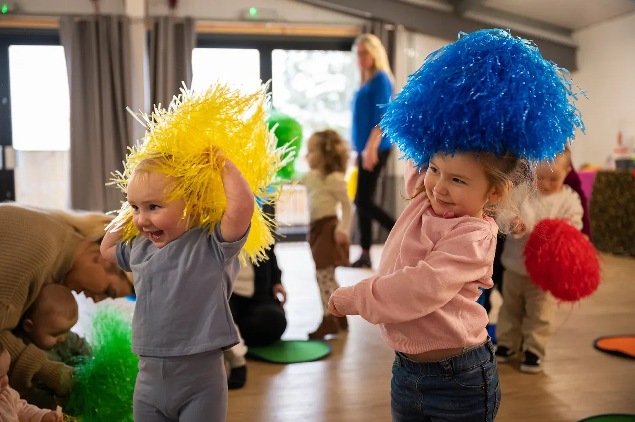 Young children wearing oversized coloured pom-pom hats dance and play together indoors during a music class.