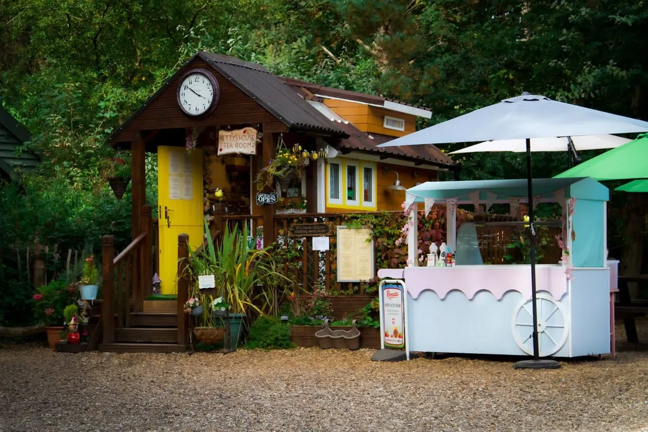 Colourful teahouse and ice cream cart nestled among trees at a leisure park, with white umbrellas providing shade.