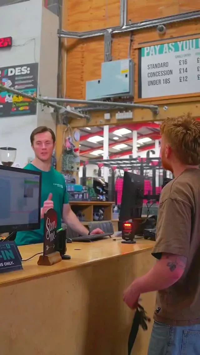 Staff member in green shirt serves customer at indoor climbing centre reception desk.