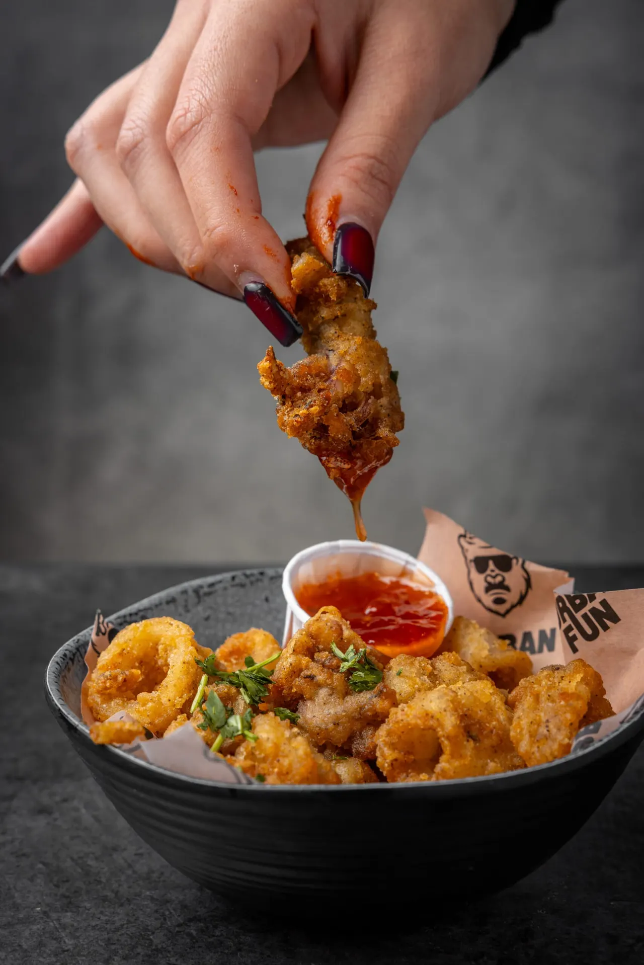 A hand holding a golden fried prawn piece dripping over a bowl of fried prawns with red dipping sauce.