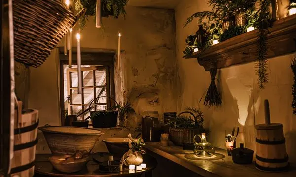 Dimly lit interior room with wooden beams, hanging plants, candles, rustic vessels, and a window, creating an atmospheric medieval or historic cottage setting.