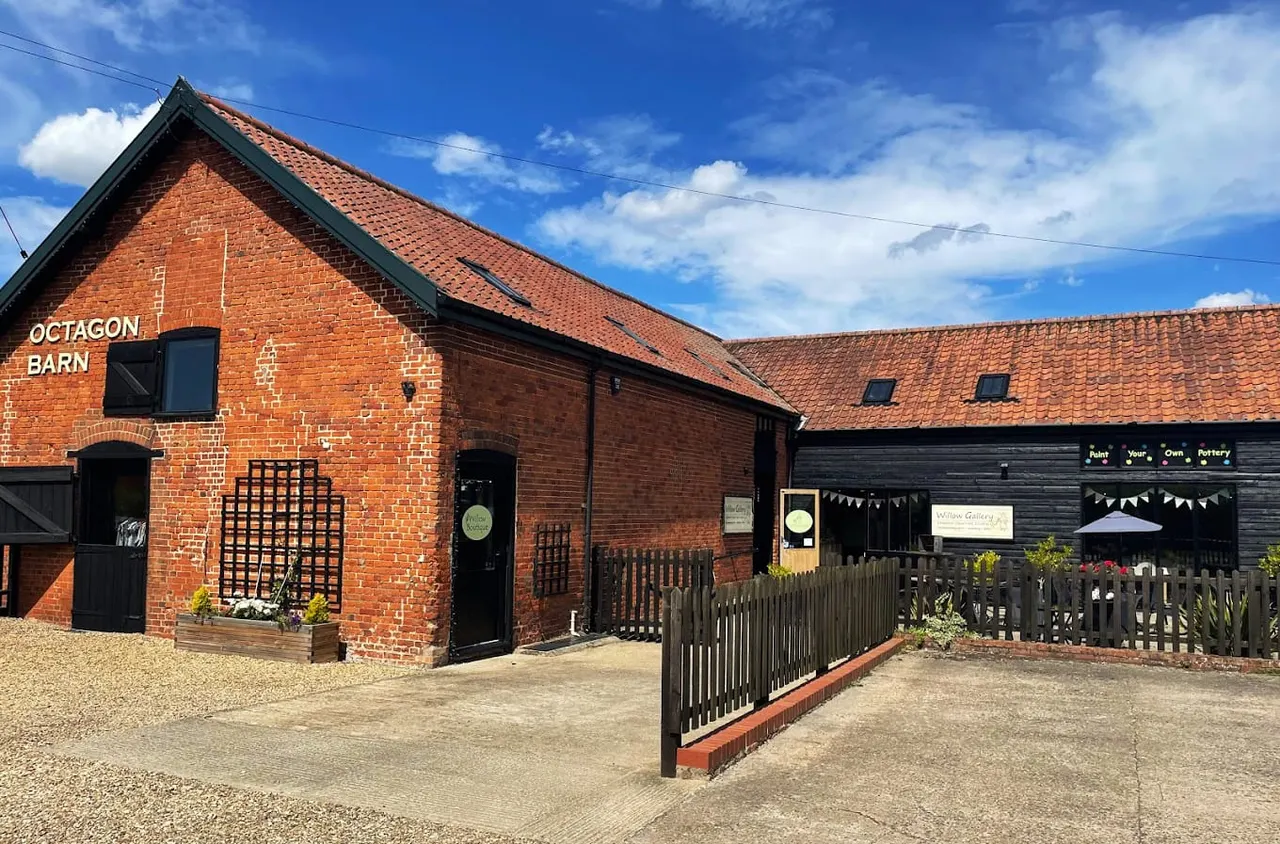 Historic red brick barn complex with black doors and windows, wooden fencing, and signage for Octagon Barn and gallery spaces under blue sky.