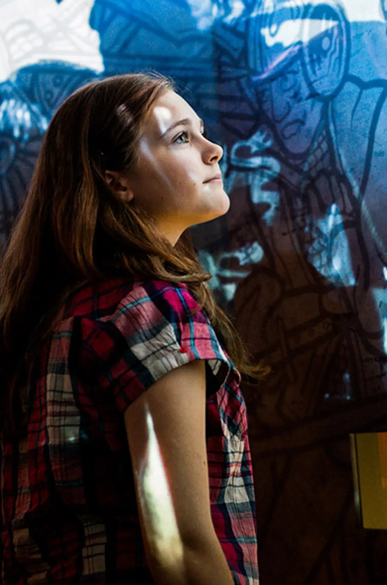 Woman in tartan shirt gazing upward at blue medieval tapestry artwork on wall.