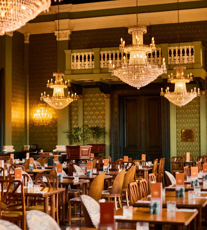 Elegant dining room with multiple crystal chandeliers, wooden tables and chairs set for service, ornate patterned wallpaper and exposed beams.