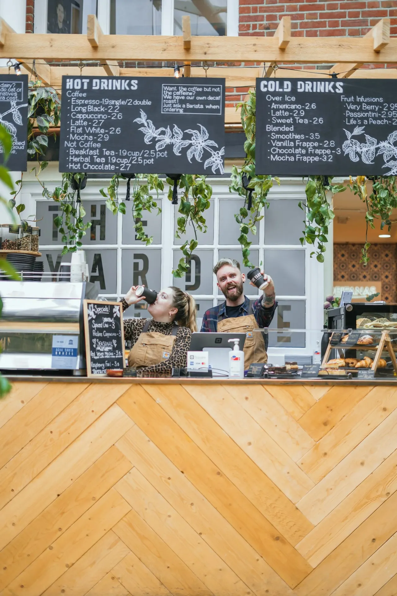 Two baristas working at a café counter beneath menu boards for hot and cold drinks, with hanging plants and pastries on display.