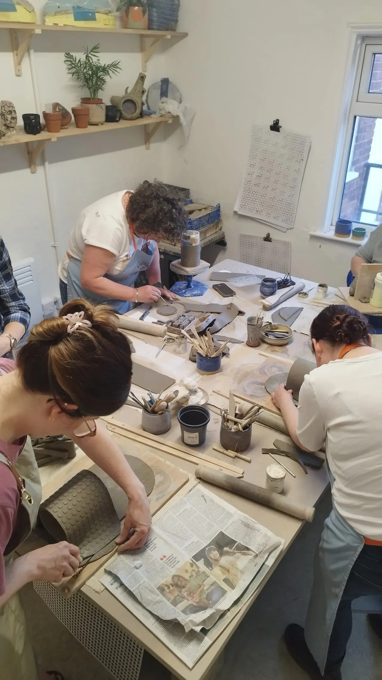 Three people working with clay at a pottery studio table surrounded by tools and materials.