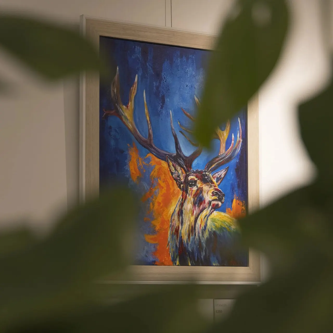 Framed painting of a stag with large antlers against a blue and orange abstract background, hung on a white wall.