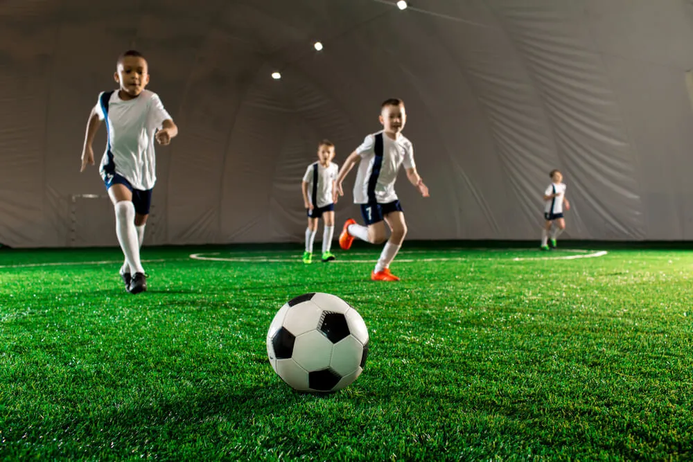 Children playing football indoors on artificial grass under bright lights.