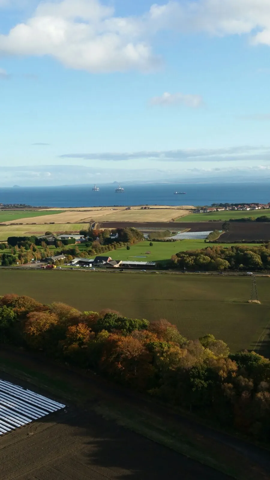 Aerial view of rolling farmland with fields, trees and a river, with sea and oil platforms visible in the distance.