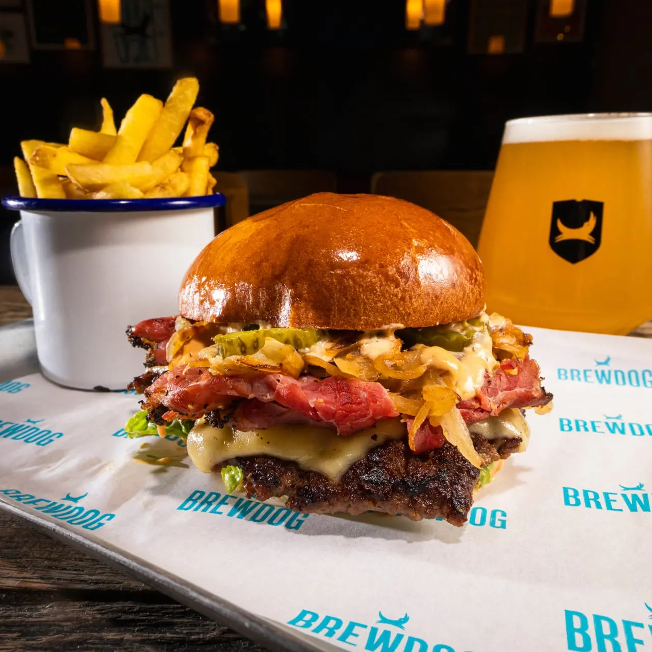 Loaded burger with bacon and fried onions served with chips and beer in a bar setting.