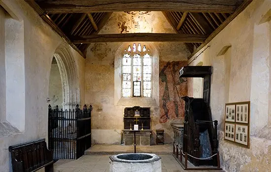 Interior of medieval chapel with stone walls, wooden roof beams, circular font in foreground, and tall arched window.