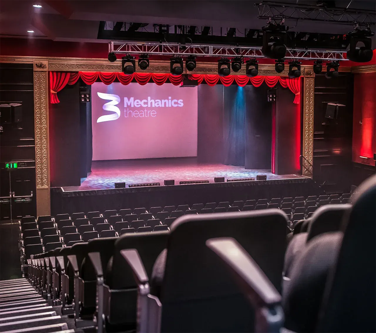 Empty theatre auditorium with red-lit stage, ornate gold proscenium arch, red swags, and rows of black seats facing the Mechanics Theatre stage.