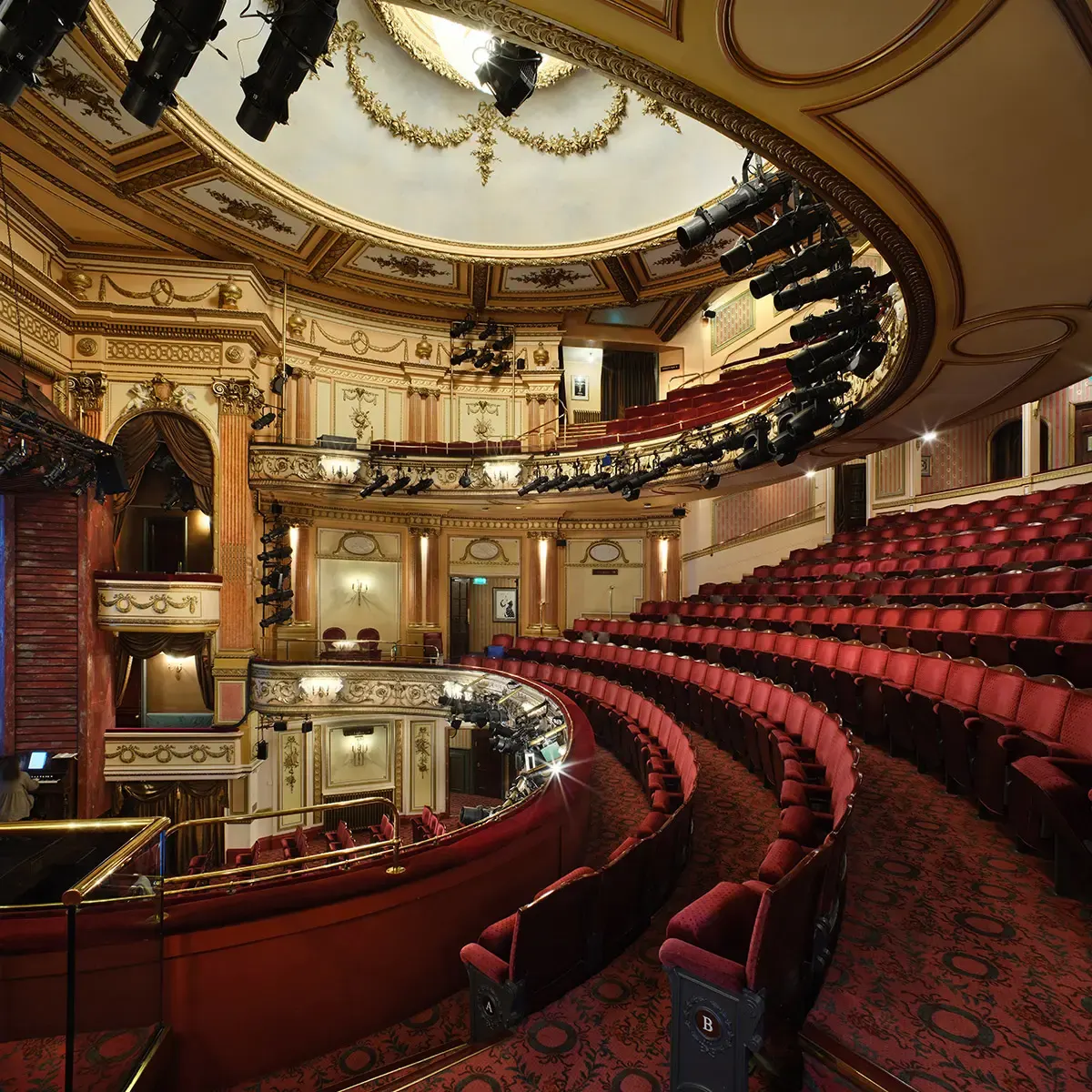 Grand theatre auditorium with red seats, ornate gold ceiling, multiple balcony levels and decorative mouldings throughout.