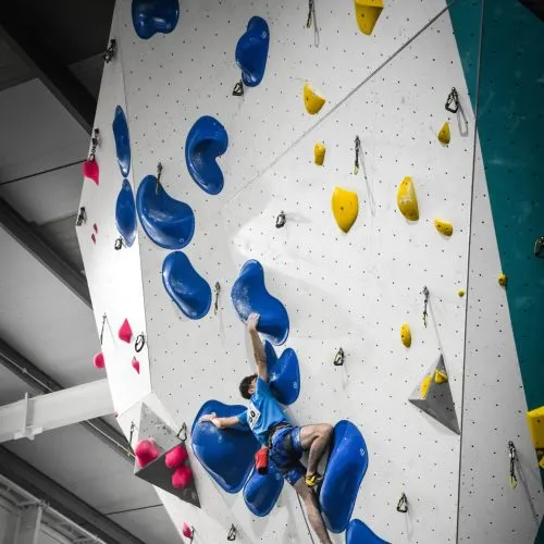 Person climbing an indoor wall with coloured handholds, viewed from below.