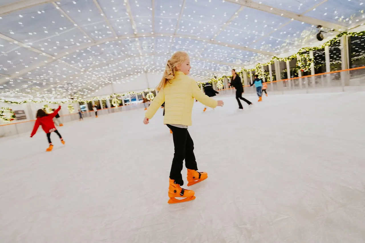 Girl in yellow jacket ice skating indoors under illuminated ceiling with other skaters in background.