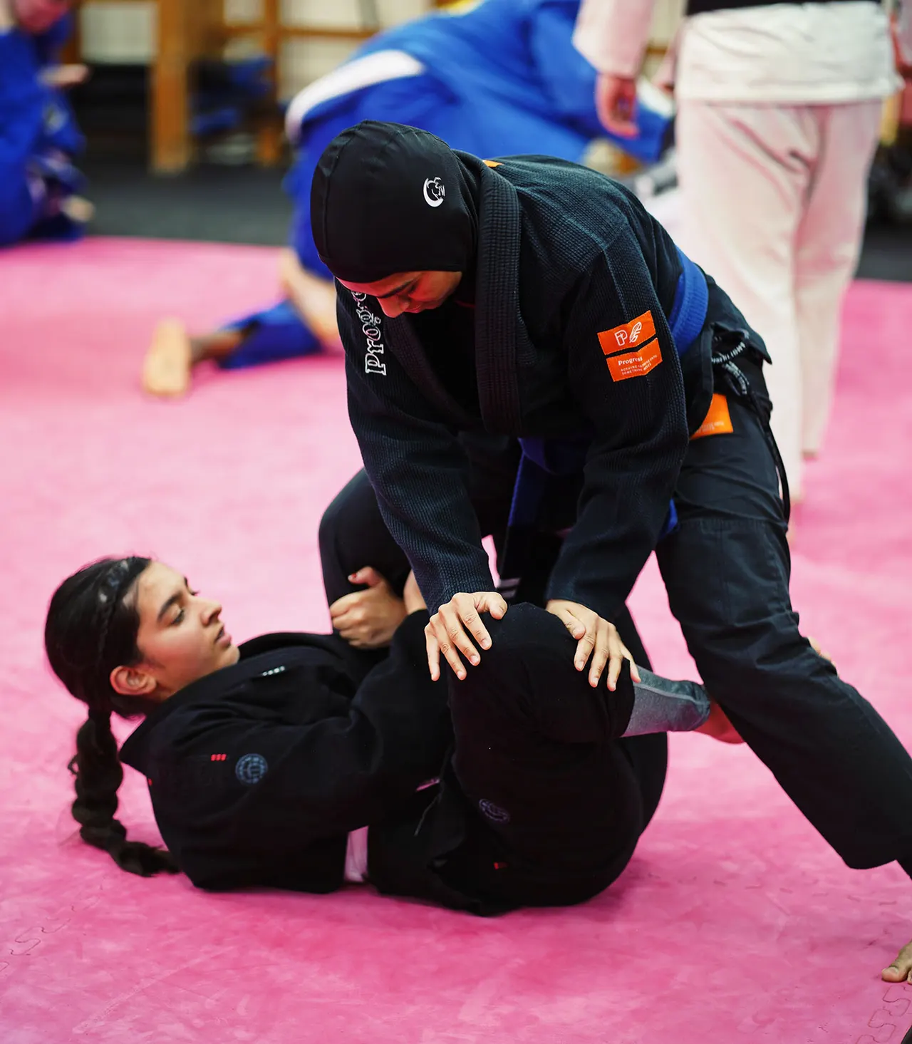 An instructor demonstrates a grappling technique on a young student during a Brazilian Jiu-Jitsu class on a pink mat.