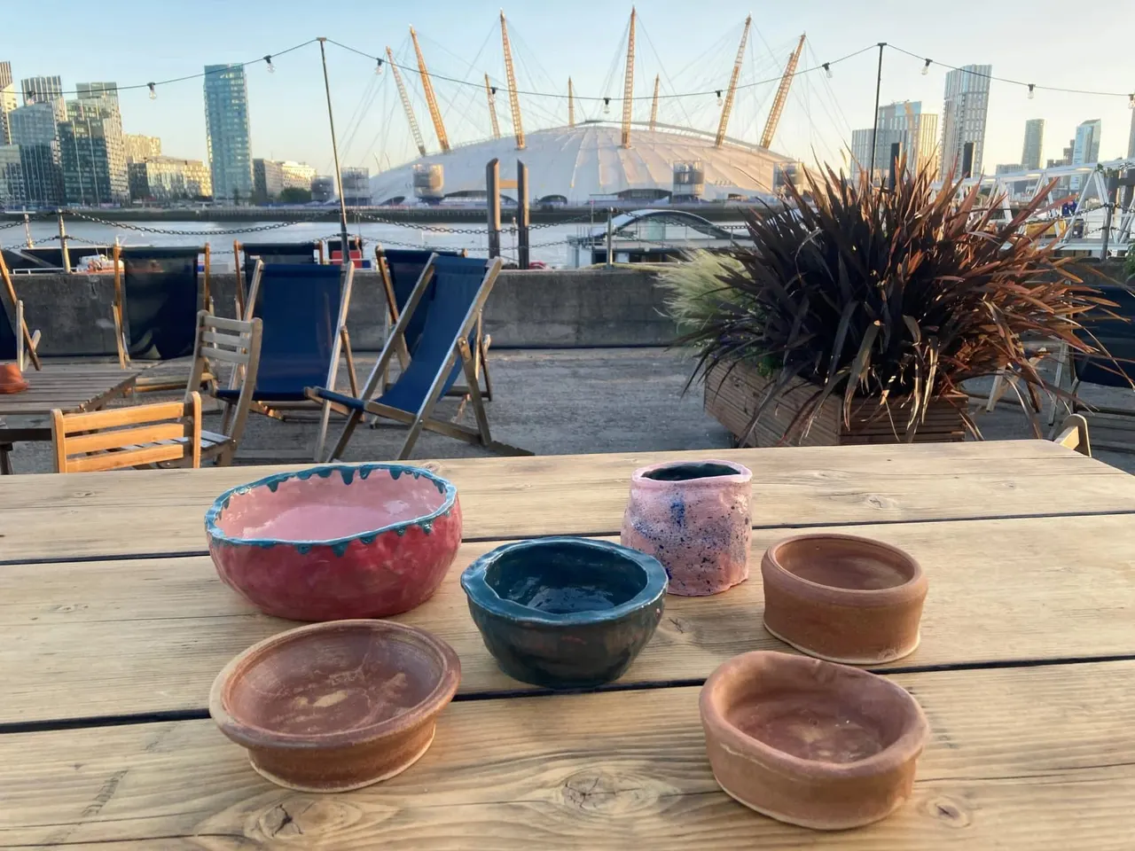 Ceramic bowls and vessels displayed on a waterside wooden table with deck chairs and a domed arena building across the water.