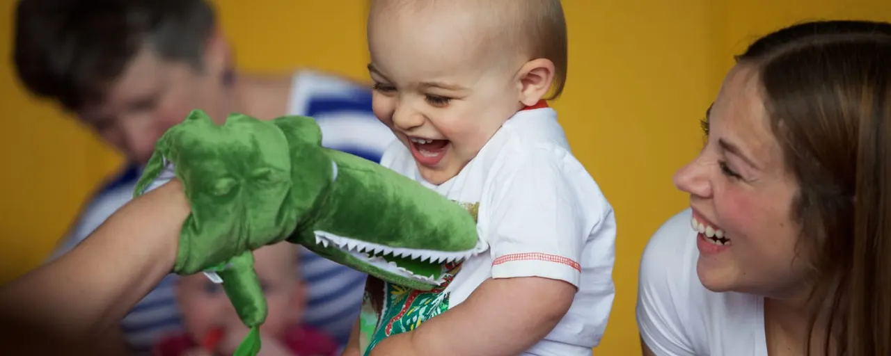 A toddler laughs with delight whilst holding a green crocodile puppet during a children's activity session.