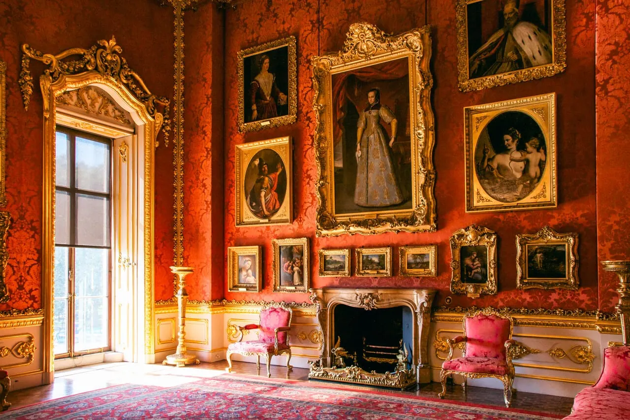 Ornate stately home interior with red damask walls, gold-framed paintings, pink velvet chairs and fireplace.