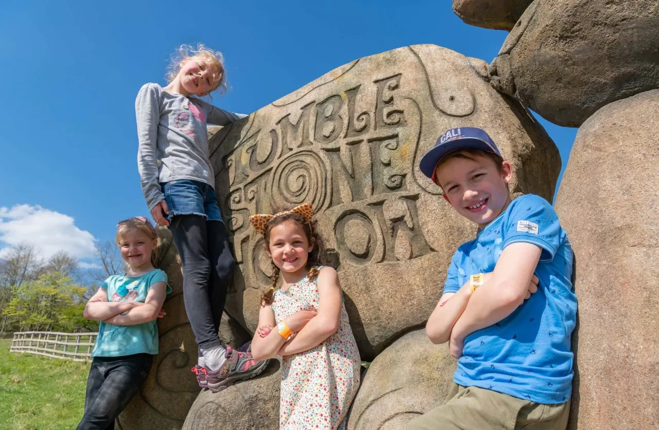 Five children posing together on and around a large carved stone boulder with text on it, under a clear blue sky.