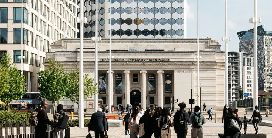 A busy public square in front of a classical stone building with columns, surrounded by modern office towers, with many people walking and standing around.