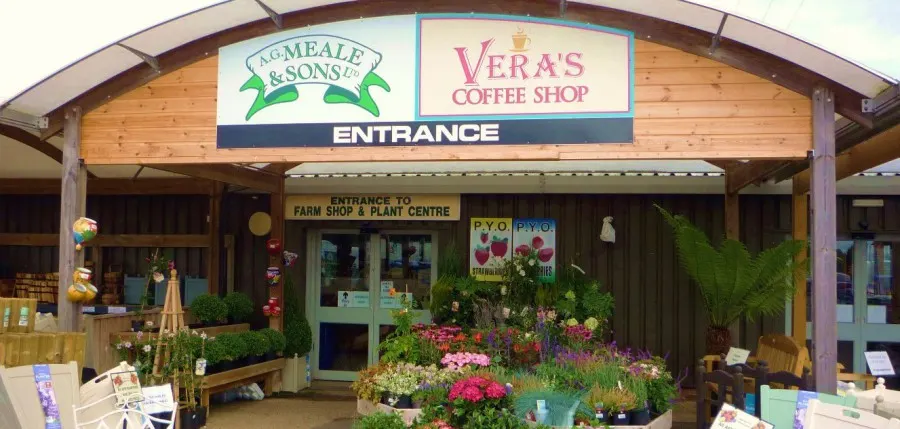The wooden-fronted entrance to a farm shop and plant centre with signage for Vera's Coffee Shop, flanked by colourful flowering plants and potted greenery.