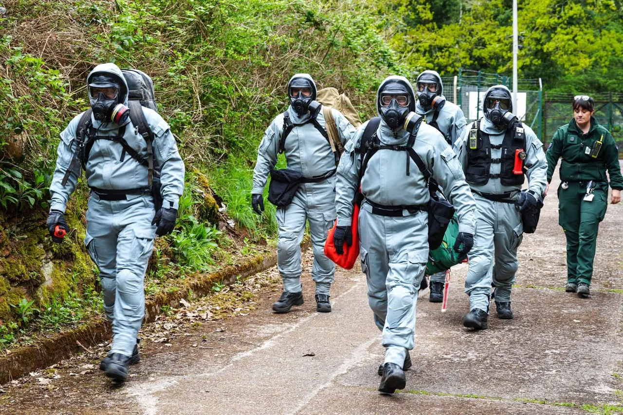 A group of people in protective suits and gas masks walk along a path lined with vegetation, accompanied by a staff member in green uniform.