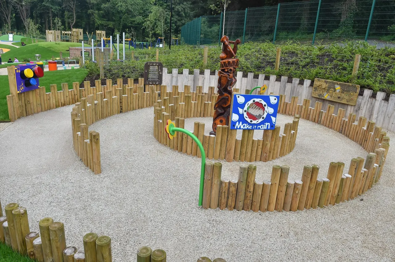 Children's outdoor play area with wooden post maze, green rope feature, and carved wooden animals on gravel surface.