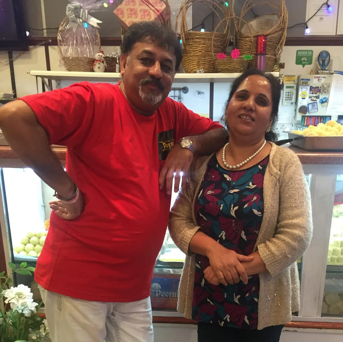 Two people posing together inside a shop with decorative baskets and shelves behind them.