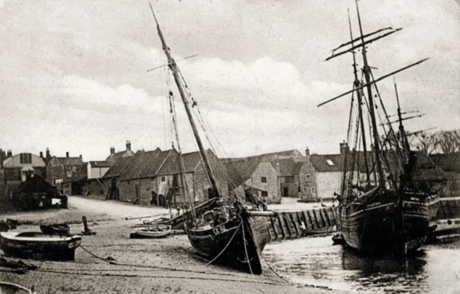 Historic black and white photograph of moored sailing vessels at low tide in a coastal village harbour.
