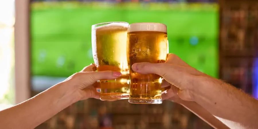 Two people clinking pint glasses of beer in front of a television screen showing sport.