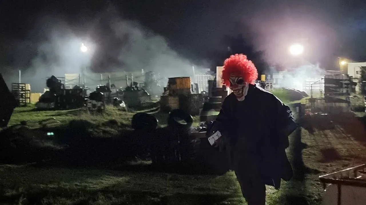 Person wearing red clown wig and dark clothing at night-time laser tag arena with illuminated obstacles and buildings.