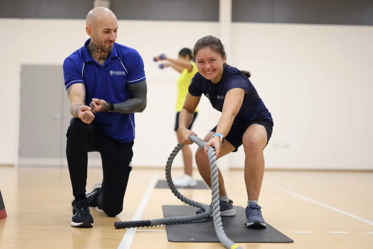 Instructor and participant performing battle rope exercise in indoor sports hall with other exercisers in background.