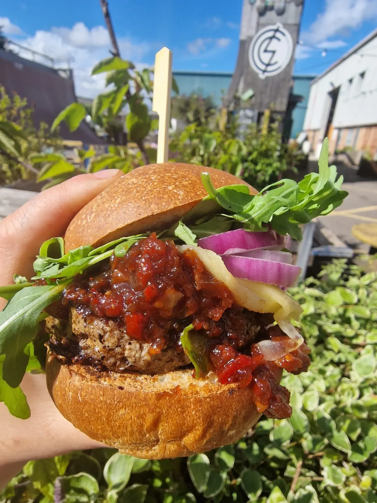 Hand holding a burger topped with sauce, fresh greens and purple onion, with a sunny garden venue visible behind.