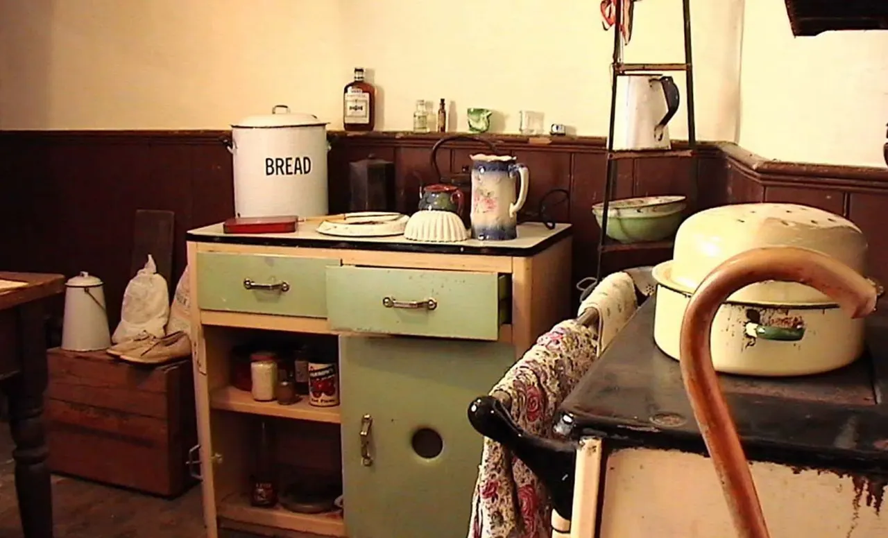 A recreated vintage kitchen interior displaying early 20th-century cooking equipment, furniture and domestic items arranged on shelves and surfaces.