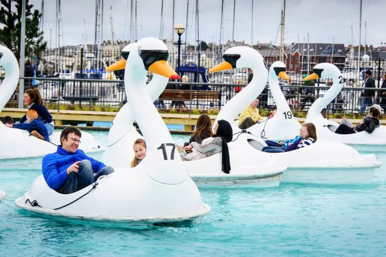 Children riding in giant swan-shaped pedalos on a shallow pool at a seaside leisure park.