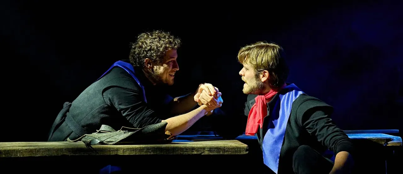 Two male actors in period costume shake hands across a wooden table during a stage performance.