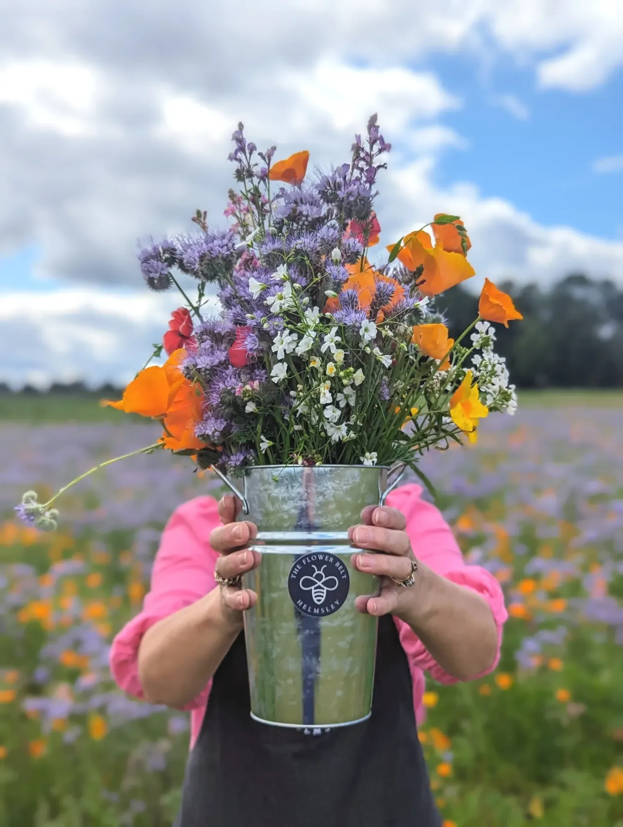 Person in pink jacket holding a vase of wildflowers in a blooming field of purple and orange flowers.