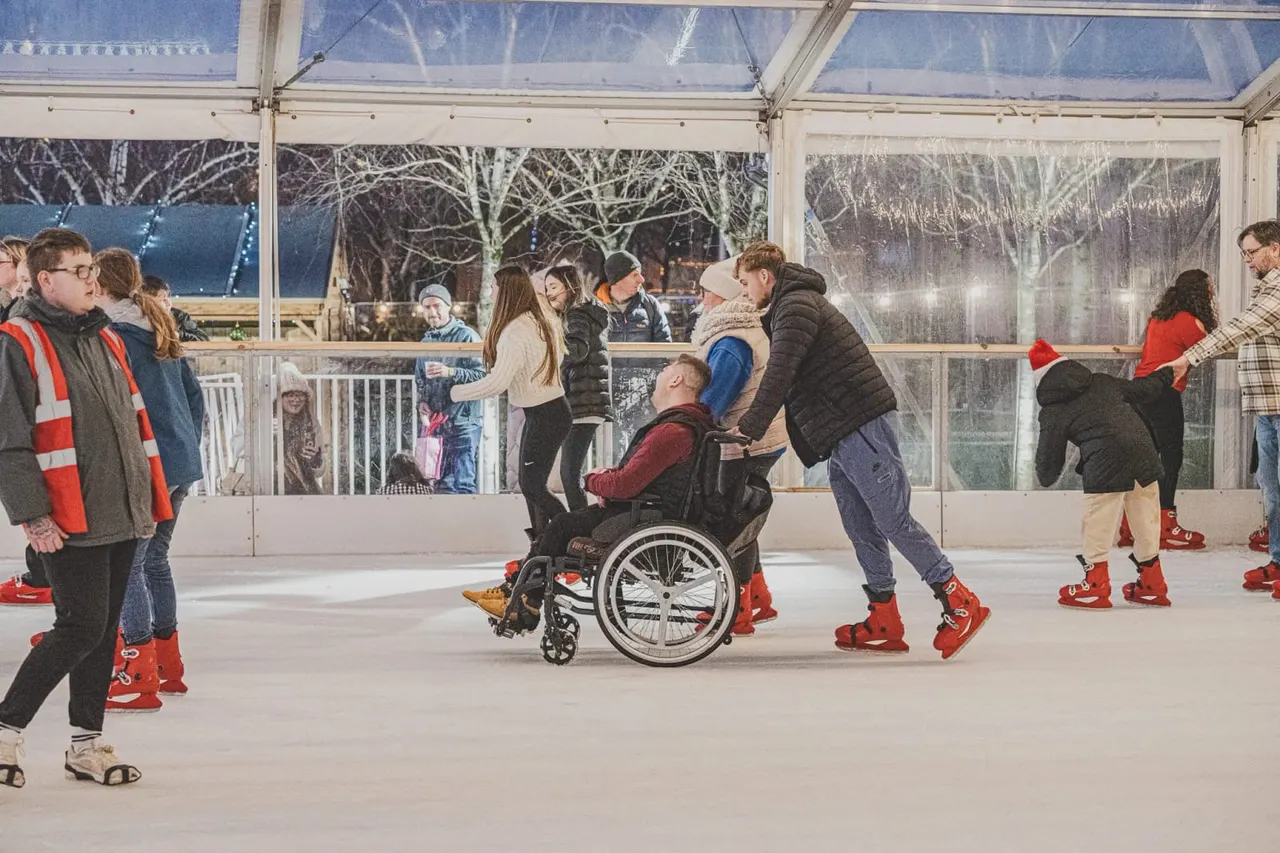 Group of people ice skating indoors in a covered rink, including a person in a wheelchair being assisted by others.