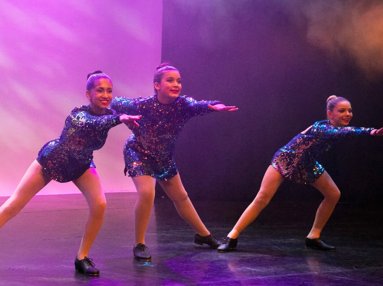 Three young dancers in matching sequined costumes performing a choreographed routine on stage under purple and pink lighting.