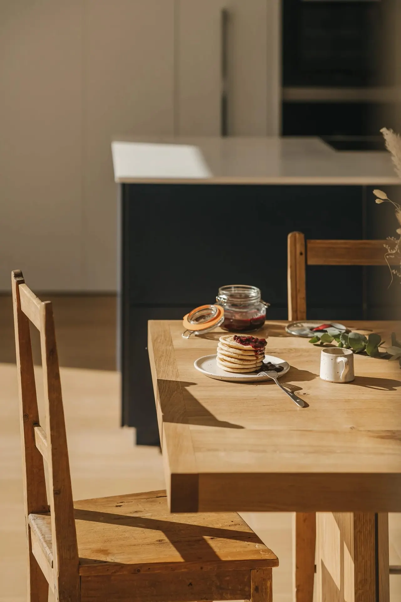 Wooden table set with stacked pancakes, jam jar and mug in a bright dining space with wooden chairs.