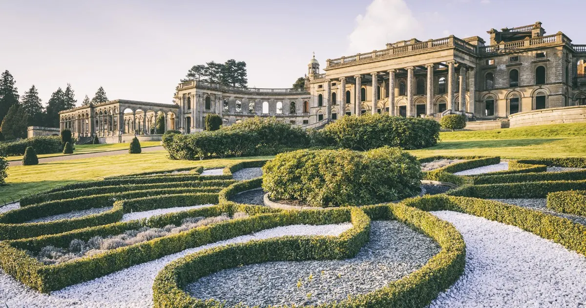 Grand stately home with classical architecture viewed across formal gardens featuring geometric hedge patterns and gravel pathways.
