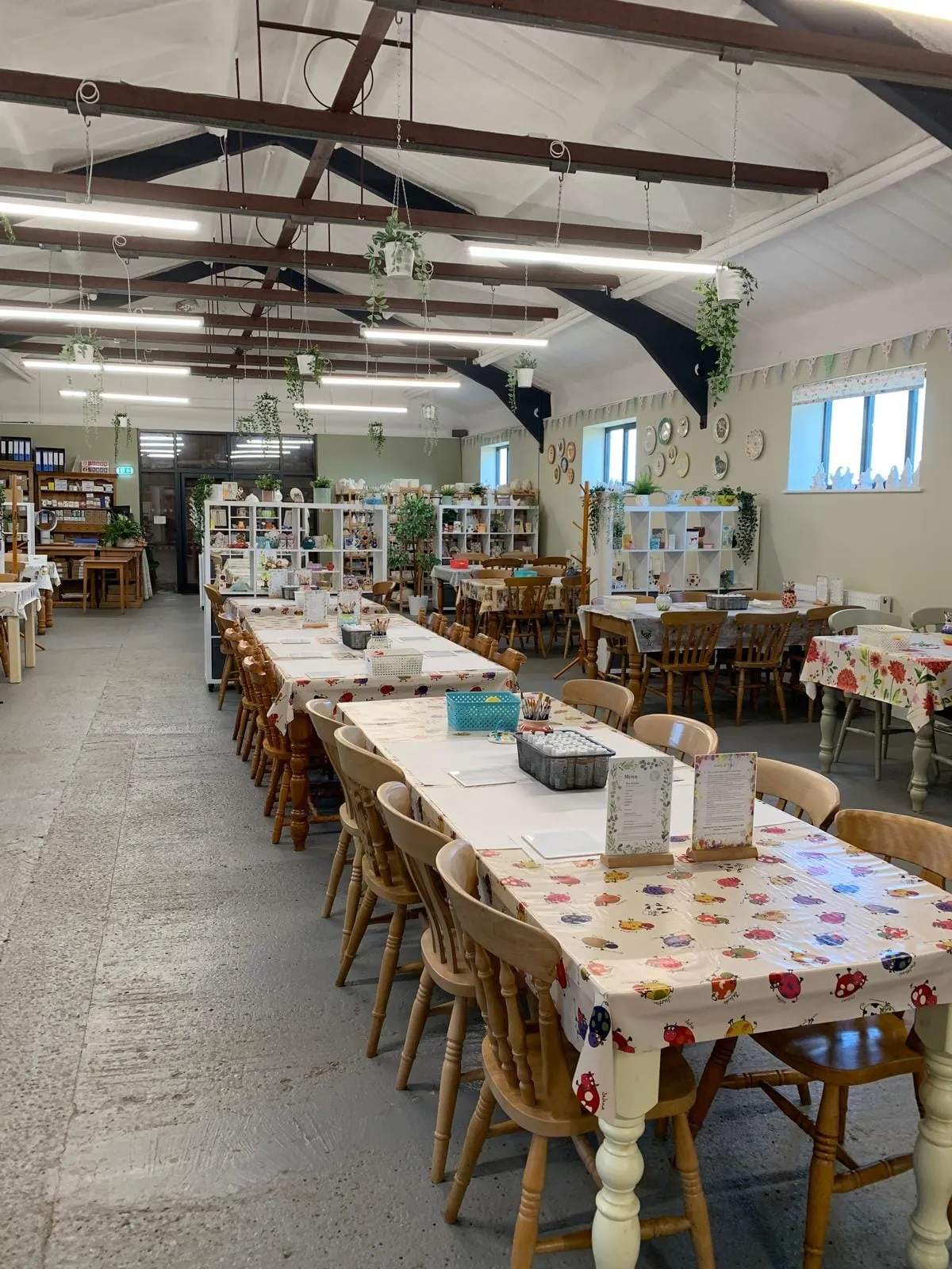 A spacious creative studio with multiple tables set up for pottery or craft activities, wooden chairs, shelving units, hanging plants, and natural light from large windows.