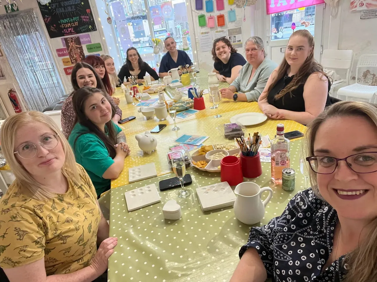 A group of people seated around a yellow polka-dot table during a paint-and-pottery workshop, with art supplies and refreshments.