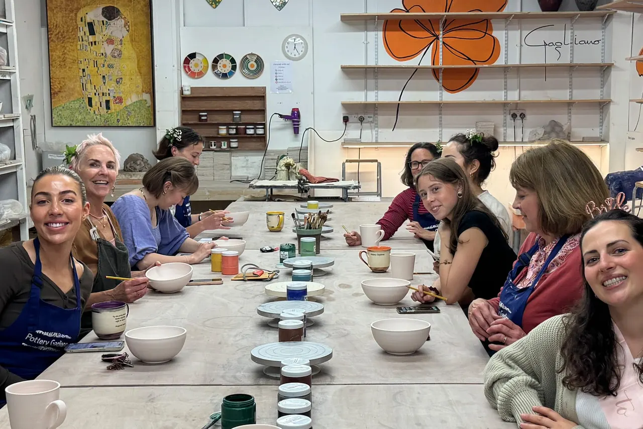 A group of smiling visitors sit at a long pottery studio table with ceramic bowls and painting supplies during a workshop.