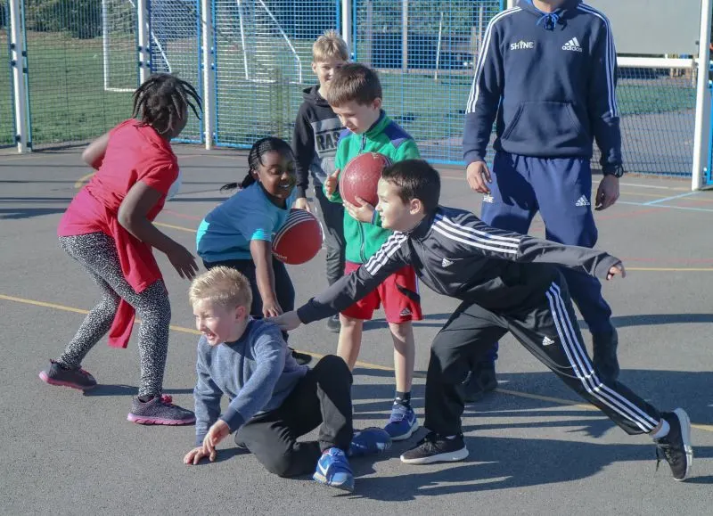 Children playing netball and football with instructors on an outdoor sports court.