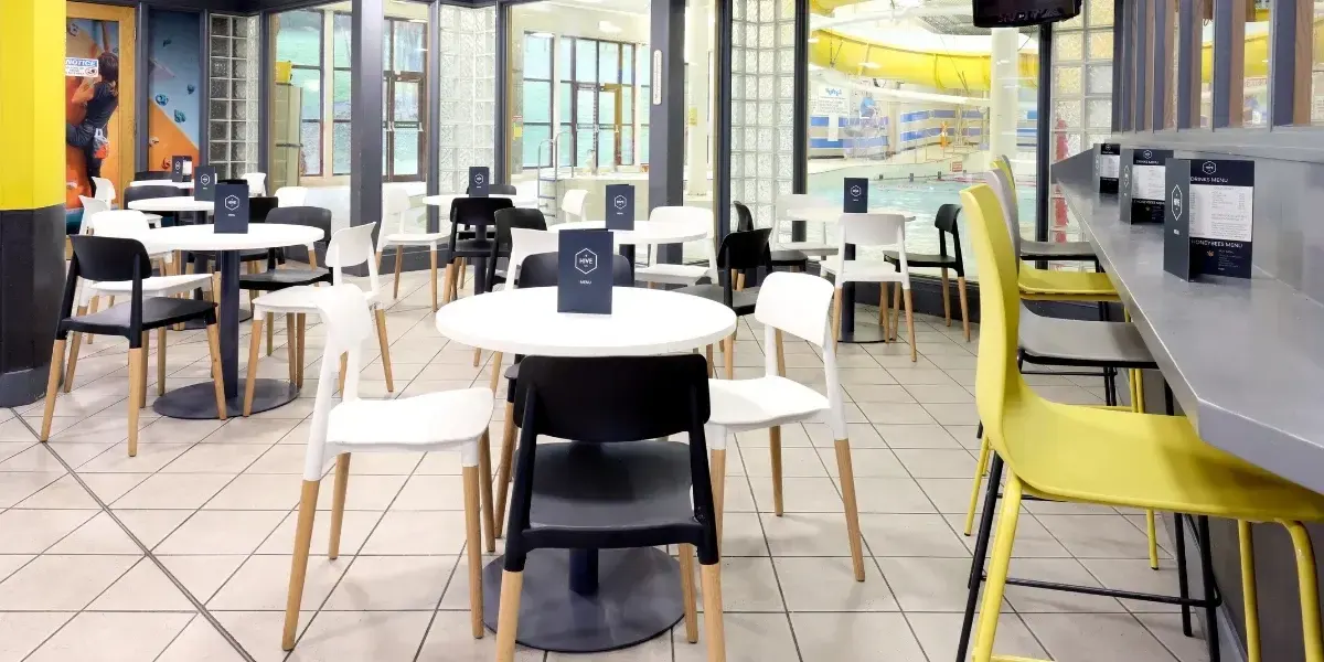 Modern café interior with white tables, black and yellow chairs, overlooking an indoor swimming pool through large windows.