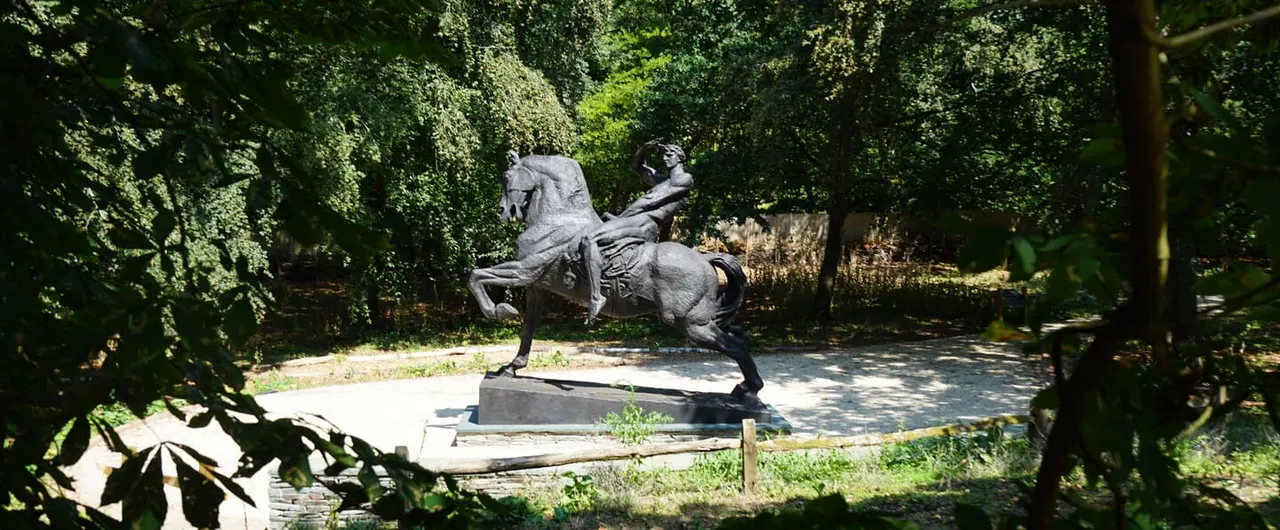 Stone sculpture of a rearing horse with rider in a wooded garden clearing, surrounded by dense green foliage.